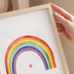 Hand hält einen Poster Regenbogen A3, A4 & A5 in einem hellen Holzrahmen, zeigt einen Aquarell-Regenbogen über einem kleinen roten Herz auf weißem Hintergrund.