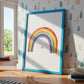 Poster Regenbogen A3, A4 & A5 im blauen Rahmen, zeigt Aquarell-Regenbogen mit Herz. Ideal für Kinderzimmer, auf hellem Holzboden mit Spielzeug und Büchern.