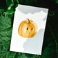 Postkarten Set Kürbis A6 zeigt eine Karte mit einem lächelnden Kürbis auf weißem Papier, platziert auf dunklen Blättern. Ideal für Herbstgrüße.