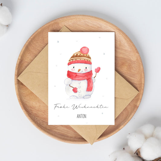 Personalisierte Weihnachtskarte Schneemann A6, zeigt handgemalten Schneemann mit Herz-Stöcken und Frohe Weihnachten Anton. Liegt auf rundem Holztablett mit Umschlag darunter.