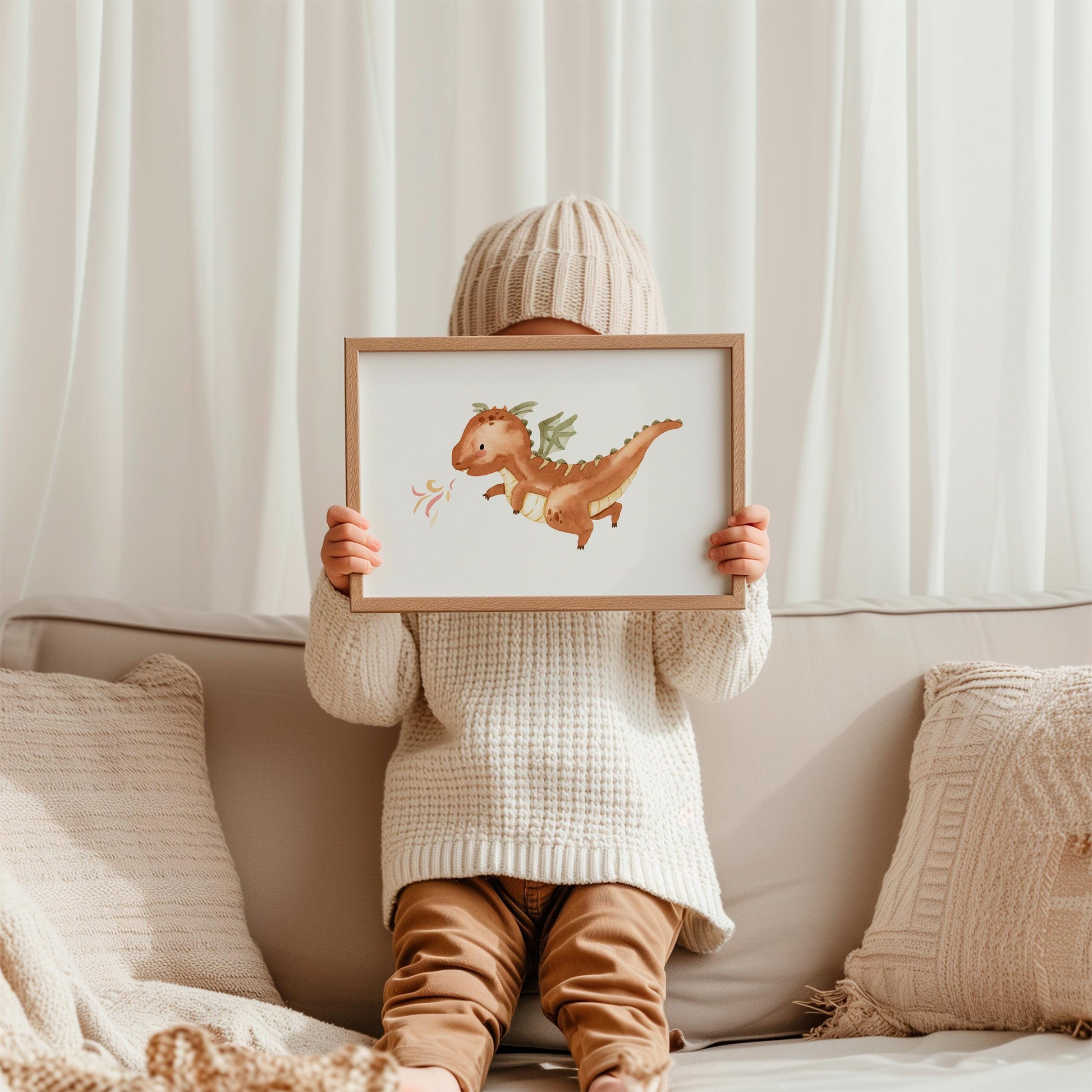 Kind hält gerahmtes Poster Feuerdrache A3 & A4 mit braunem Drachen, grünen Flügeln, gemalt in Aquarellstil, auf einer Couch sitzend, hoch.
