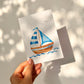 Postkarte Segelboot A6 zeigt ein Aquarell-Segelboot mit blauen Streifen, schwebend über sanften Wellen, auf hochwertigem, beschreibbarem Naturpapier gedruckt.