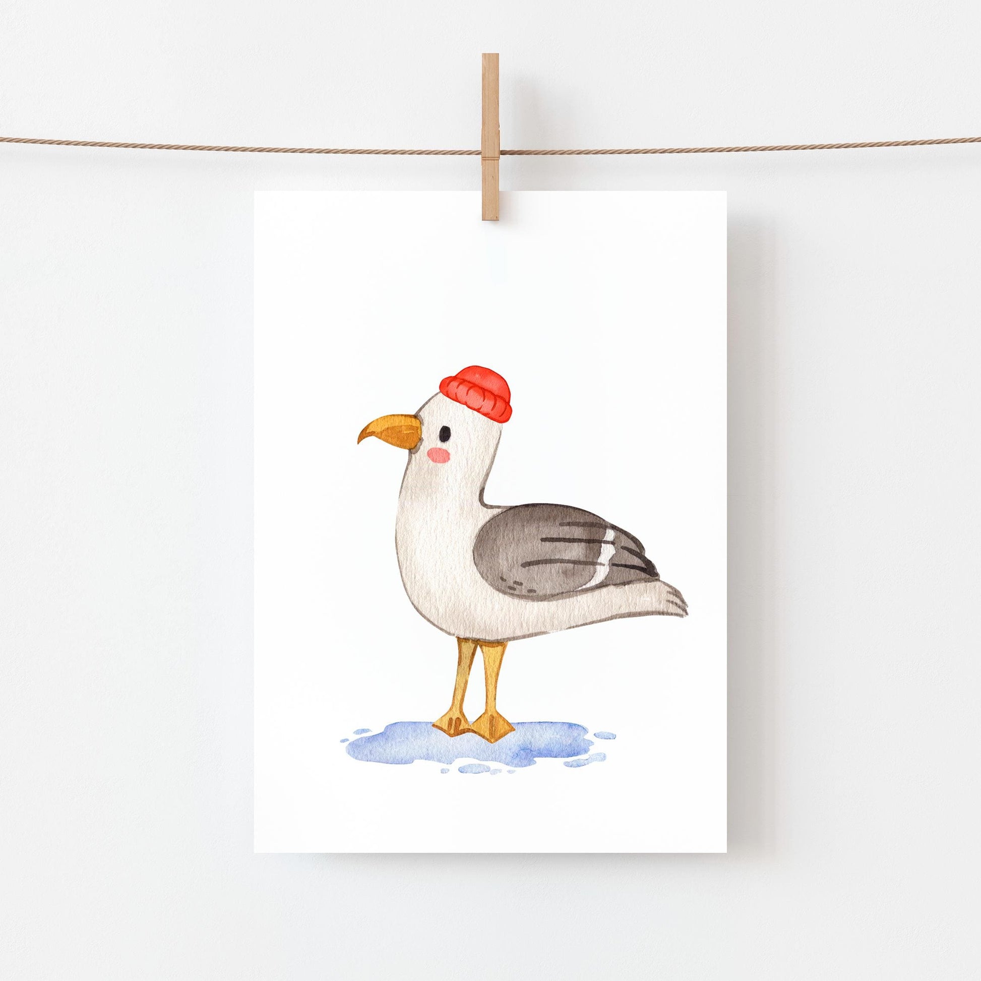 Postkarte Möwe A6 zeigt eine Möwe im Aquarellstil auf Naturpapier, mit gelbem Schnabel, grauen Flügeln und roter Mütze, aufgehängt an einer Kordel.