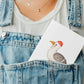 Postkarte Möwe A6 steckt im Latz einer Jeanslatzhose, über weißem T-Shirt, zeigt illustrierten Vogel mit roter Mütze. Goldene Halskette sichtbar.