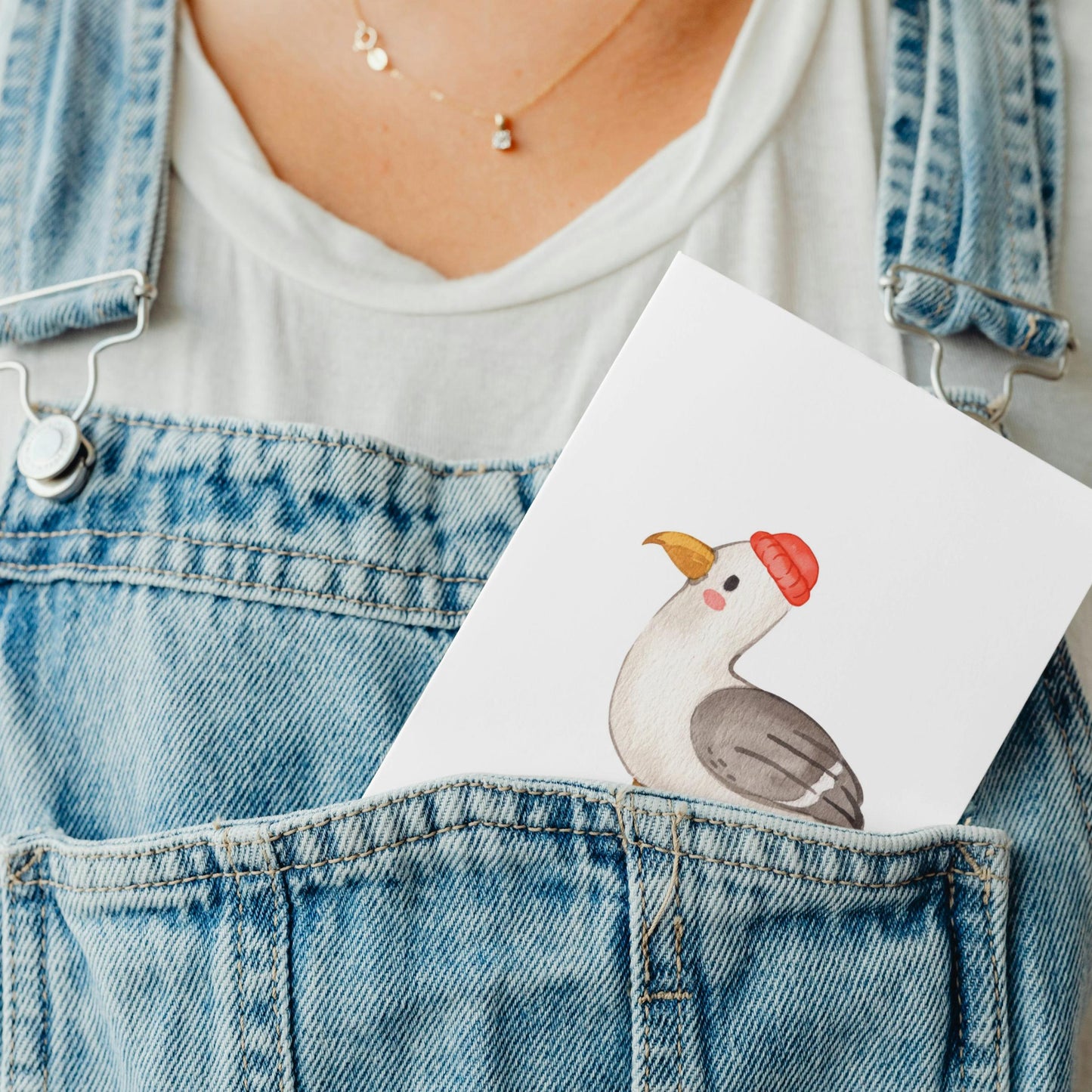 Postkarte Möwe A6 steckt im Latz einer Jeanslatzhose, über weißem T-Shirt, zeigt illustrierten Vogel mit roter Mütze. Goldene Halskette sichtbar.