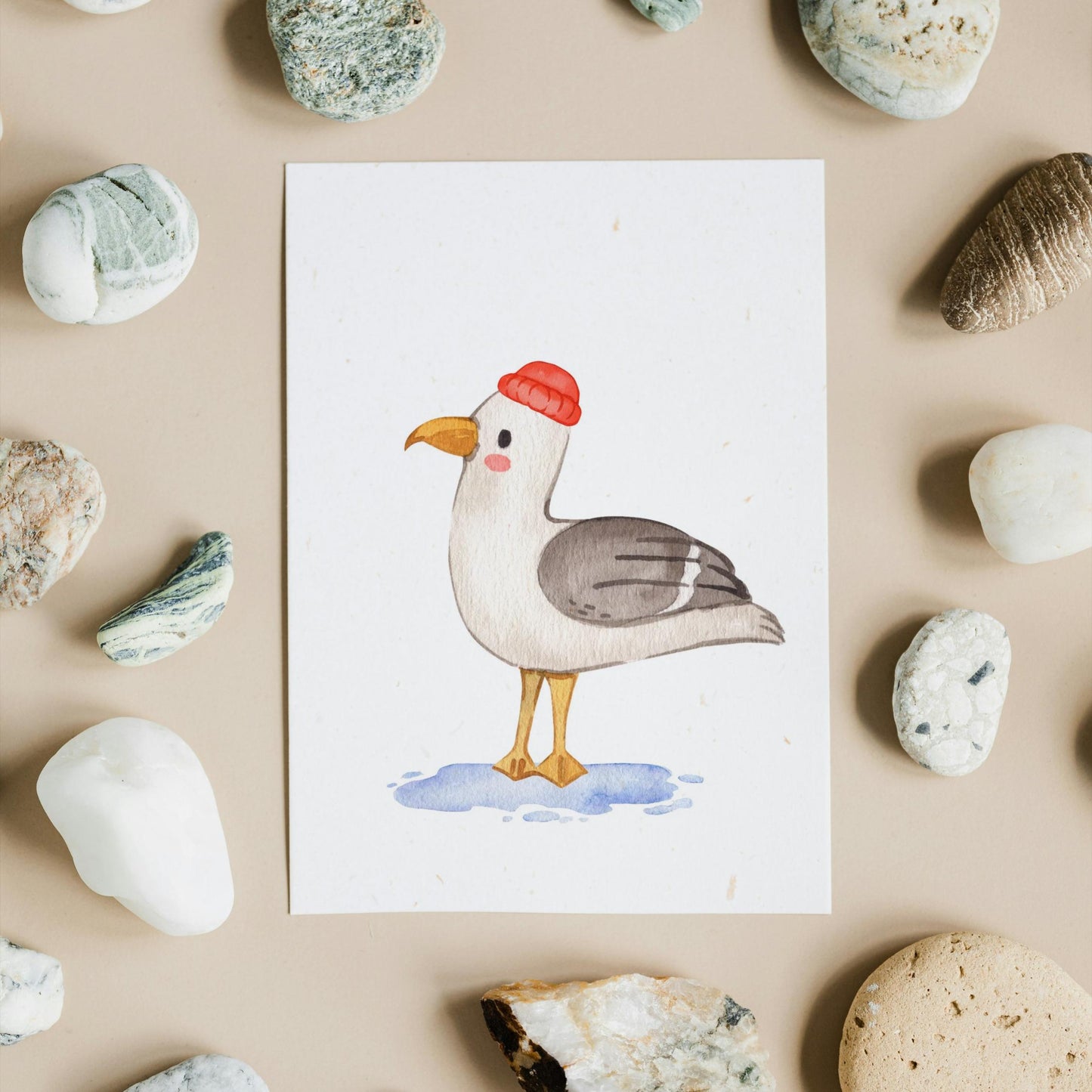 Postkarte Möwe A6 mit Aquarell-Motiv einer Möwe in roter Mütze, umgeben von Steinen. Gedruckt auf hochwertigem, mattem Naturpapier, ideal für Grüße oder Dekoration.