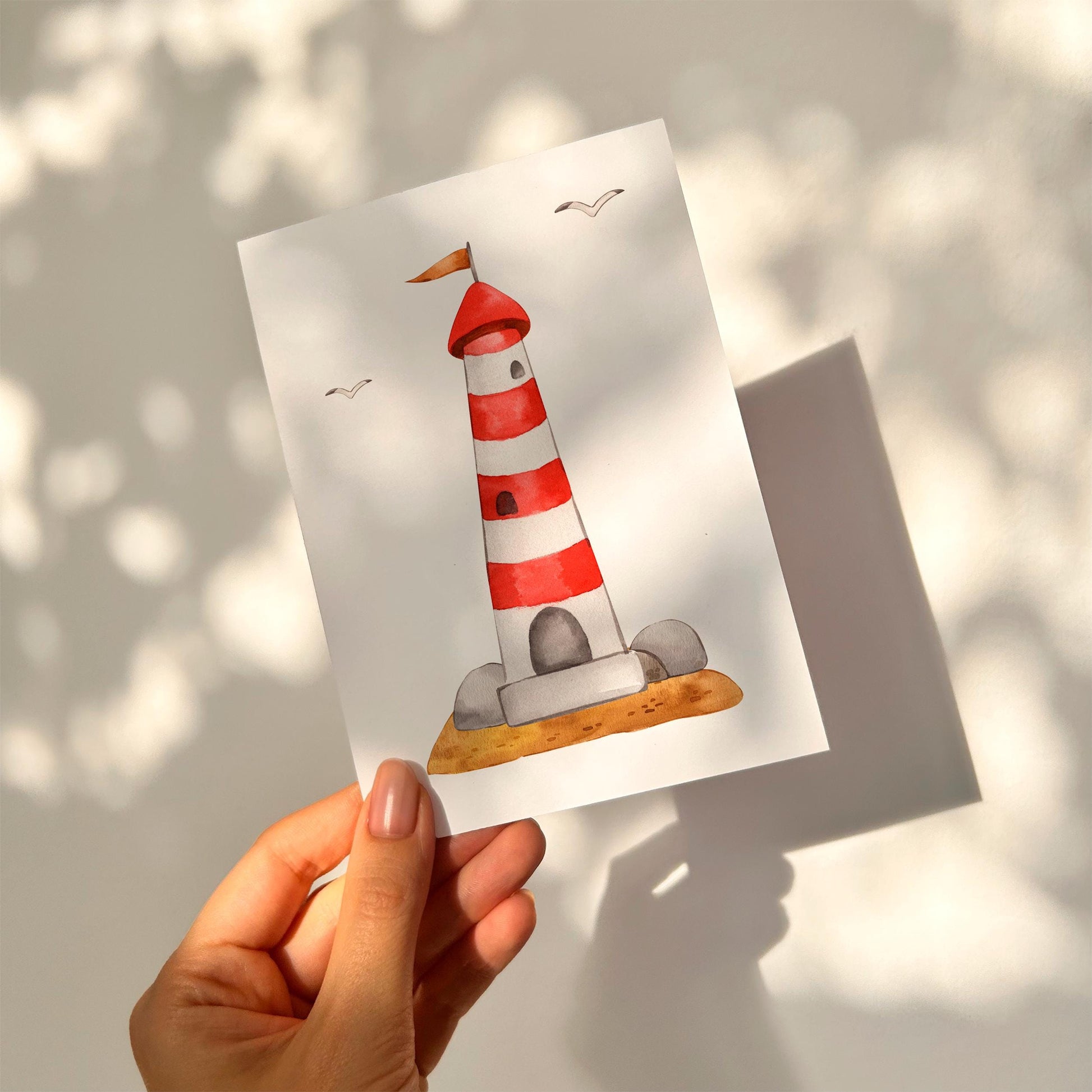 Person hält die Postkarte Leuchtturm A6 mit rotem Leuchtturm und Möwen, ideale maritime Dekoration oder Grußkarte, auf hochwertigem, stabilem Naturpapier gedruckt.